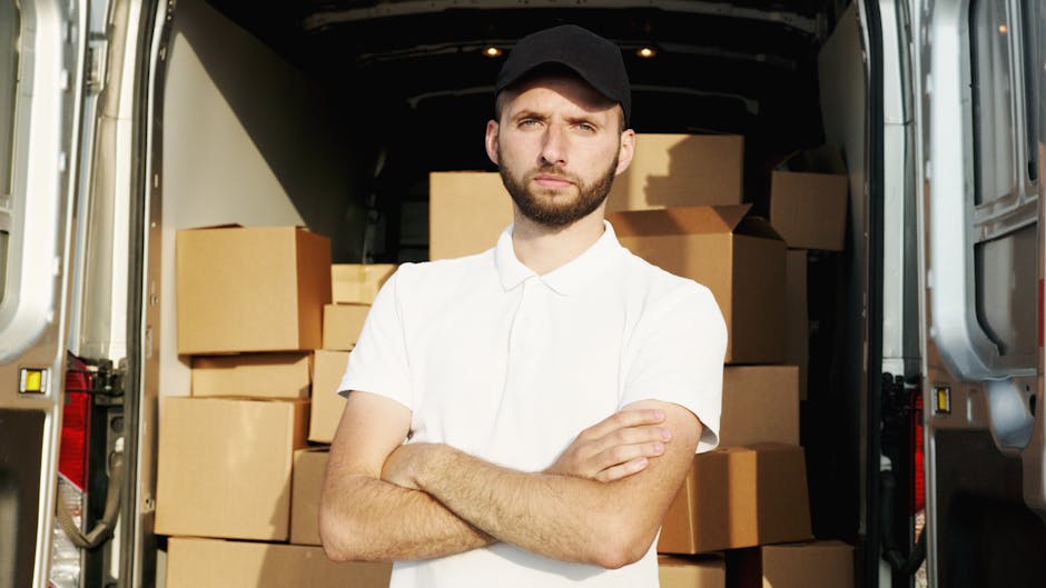 A man with a serious expression, wearing a white polo shirt and a black cap, stands inside the open back of a large moving van filled with various cardboard boxes, some sealed with packing tape and ot