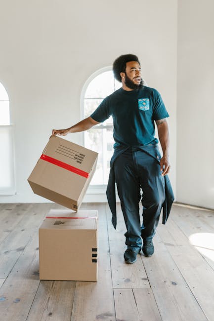 A young woman with curly black hair, wearing a white t-shirt, light blue jeans, and white sneakers, sitting on a wooden floor surrounded by numerous large cardboard boxes during a home relocation proc