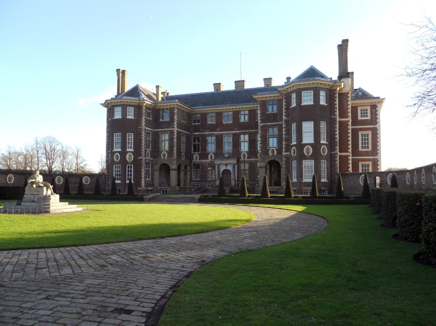 A large, multi-story brick mansion with white-framed windows and ornate architectural details is visible in the background, set within a well-maintained garden featuring a curved stone pathway. The pa