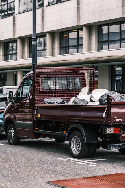 A dark red flatbed pickup truck parked on a city street in front of a modern building with large windows. The vehicle's cargo area is filled with various belongings prepared for home relocation, inclu