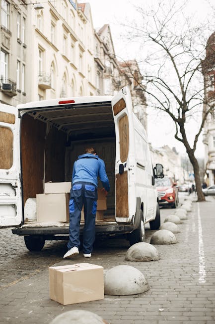 A dark red flatbed pickup truck parked on a city street in front of a modern building with large windows. The vehicle's cargo area is filled with various belongings prepared for home relocation, inclu